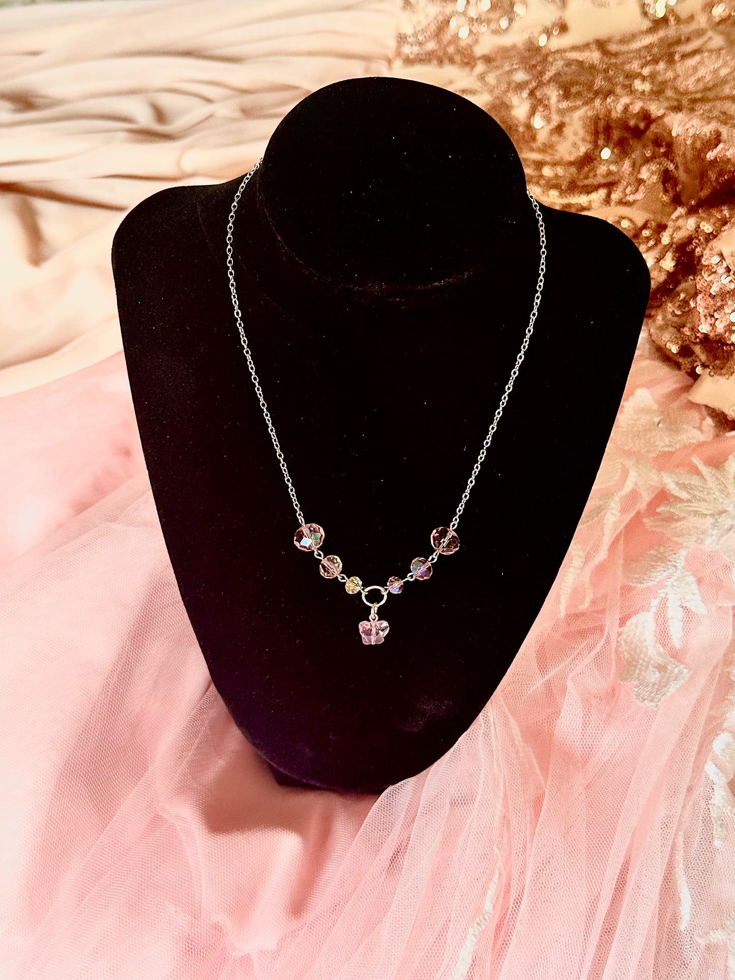 The Aurora Grace Necklace displayed on a black mannequin, featuring sparkling pink crystals arranged delicately on a gold chain against a soft pink background.