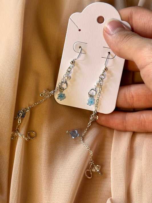 A pair of delicate, handcrafted earrings displayed on a beige card holder, featuring silver chains adorned with small blue crystals and tiny floral charms, hanging gracefully against a soft, peachy fabric background.